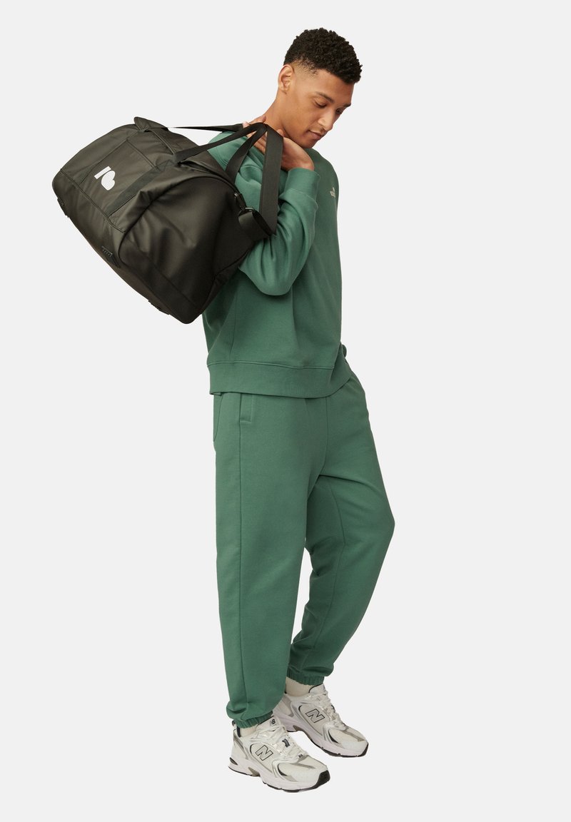 Sweatshirt en coton vert et joggers assortis, portés avec des baskets blanches. Sac de sport noir avec logo, porté à l'épaule. Texture lisse.