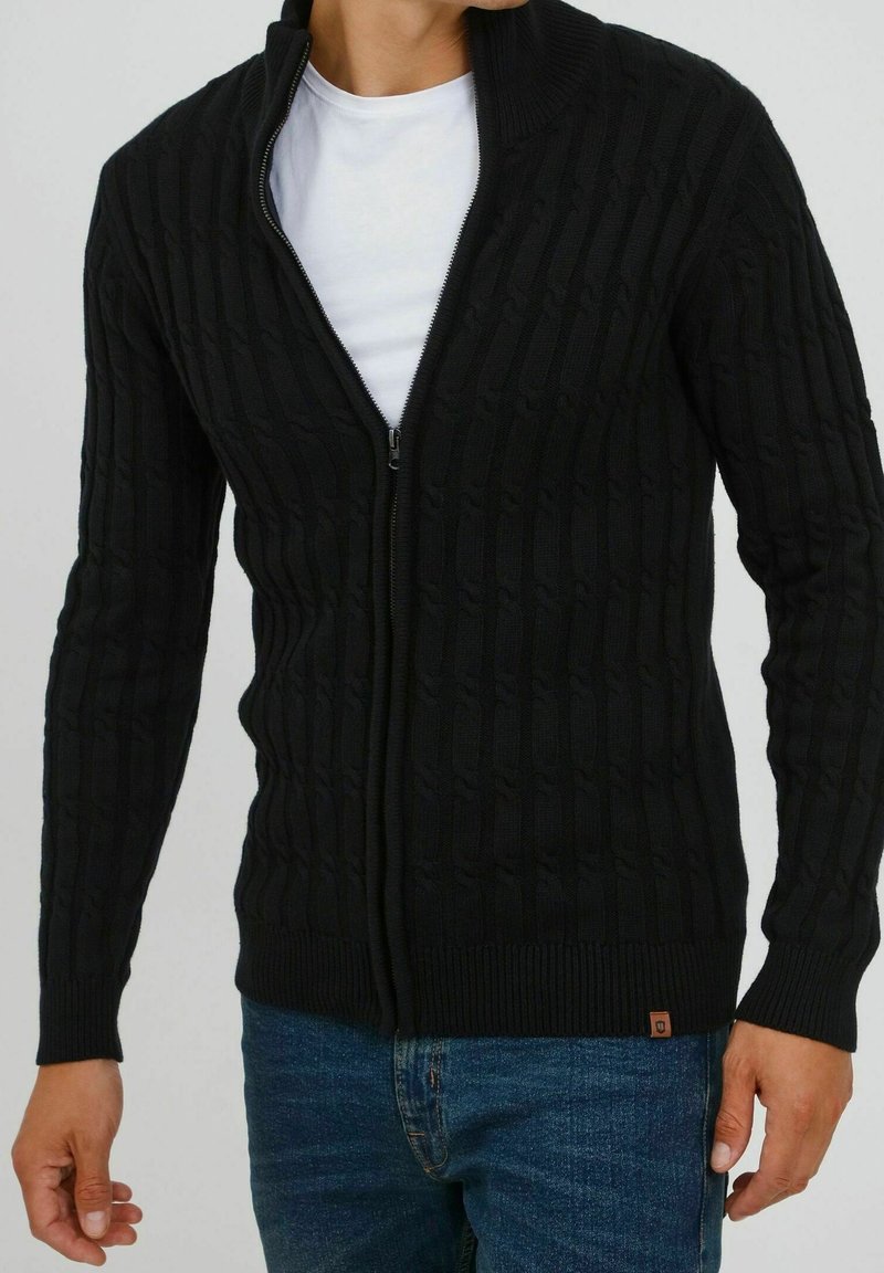 Czarny dziergany sweter zapinany na zamek z teksturowanym wzorem, wysokim kołnierzem i ściągaczami na rękawach. Noszony na białej koszuli, zestawiony z niebieskimi jeansami.
