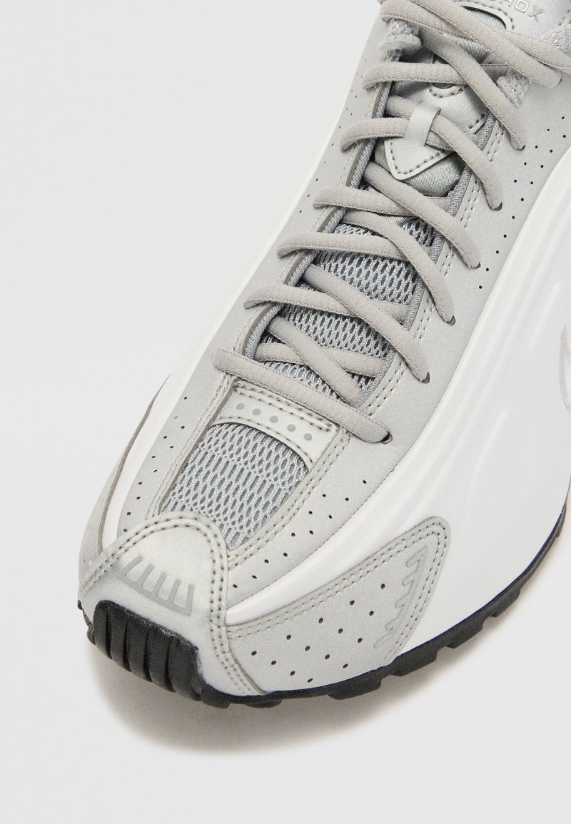 Zapatilla deportiva gris con parte superior de malla, detalles perforados y una media suela blanca. La suela exterior es negra con un patrón texturizado para mayor agarre.