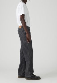 Muž stojící v profilu, oblečený v bílém tričku, volných černých džínách a černých šněrovacích botách, na jednobarevném šedém pozadí.