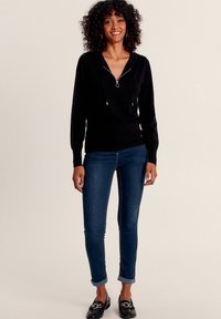 Zwarte hoodie met een ritssluiting bij de hals, gecombineerd met blauwe jeans en zwarte instappers. De hoodie heeft trekkoorden en een relaxte pasvorm.