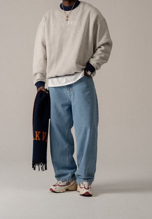 Personne portant un sweat-shirt beige, un jean bleu clair, des baskets blanches, tenant une écharpe bleu marine avec un texte orange sur fond neutre.