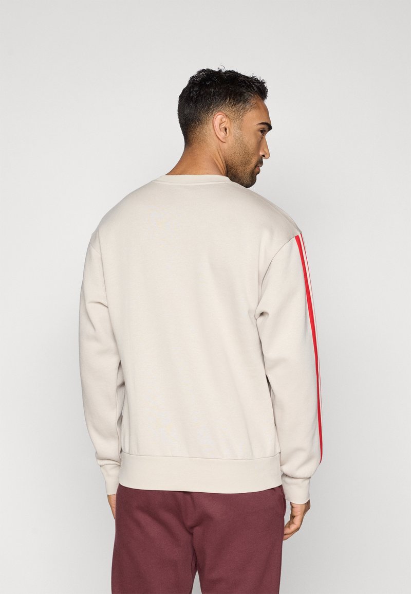 Man die zijwaarts en iets naar achter kijkt, gekleed in een beige sweatshirt met rode strepen op de mouwen en kastanjebruine broek tegen een effen achtergrond.