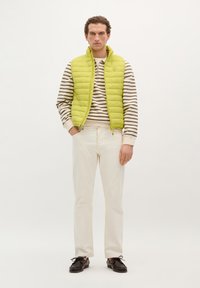 Lichtgroen gewatteerd vest over een gestreepte beige en witte sweater, gecombineerd met off-white broek en zwarte veterschoenen. Effen achtergrond.