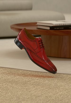 Chaussure habillée en cuir rouge à bout pointu, perforations décoratives le long des côtés, lacets noirs, et talon noir bas, flottant au-dessus d'une surface texturée.