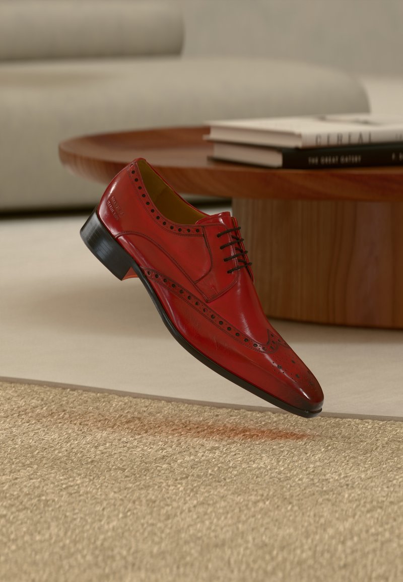 Chaussure habillée en cuir rouge à bout pointu, perforations décoratives le long des côtés, lacets noirs, et talon noir bas, flottant au-dessus d'une surface texturée.