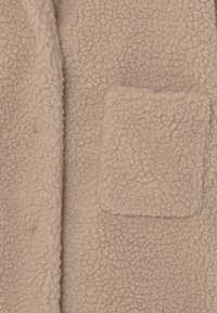 Suddig beige fleece-material med en fyrkantig ficka. Texturerad yta med ett mjukt, plysigt utseende, med en rullad krage och öppen front.