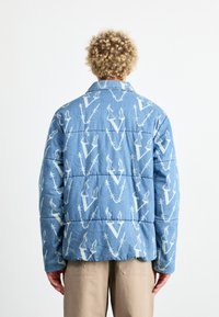 Chaqueta de mezclilla azul con textura acolchada que presenta un patrón repetido en blanco de letras y diseños. Cuello puntiagudo y mangas largas.