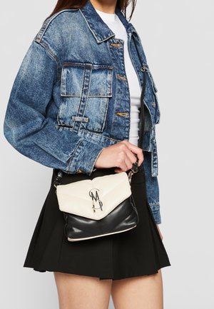 Personne portant une veste en denim bleue, un top blanc, une jupe noire, tenant un petit sac bandoulière noir et blanc avec un logo en métal sur le devant.