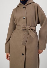 Manteau à capuche marron en matériau doux, avec ceinture nouée, gros boutons et manches longues. Texture lisse avec une coupe décontractée.