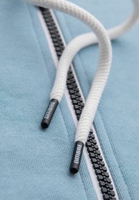 Gros plan sur des lacets blancs avec des embouts noirs portant l'inscription "DIMBRE", reposant sur une fermeture éclair en tissu bleu clair.