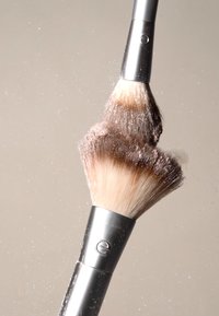 Dwa pędzle do makijażu z metalowymi, srebrnymi rączkami, z gęstymi, miękkimi włosiem w jasnobrązowym i ciemnobrązowym kolorze, tworzą chmurę cząsteczek pudru.