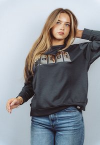 Młoda kobieta o długich blond włosach, ubrana w ciemną bluzę z nadrukiem oraz niebieskie dżinsy, pozująca z jedną ręką założoną za głowę.