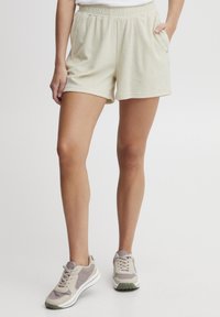 Shorts beige clair en tissu doux et texturé, avec une taille élastique et des poches latérales. Assortis à des baskets grises claires.
