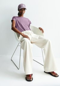 Purple short-sleeve t-shirt, beige wide-leg pants, black sandals with red accents, seated in a white chair against a light background.