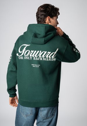 Grüner Hoodie mit Kängurutasche, auf der Rückseite mit weißem Schriftzug "Forward OR ONLY BACKWARDS", kombiniert mit hellblauen Jeans.