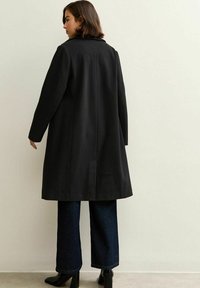 Svart ullblandning kappa med klassisk krage, långa ärmar och rak silhuett, med en bakre sprund för enkel rörelse.
