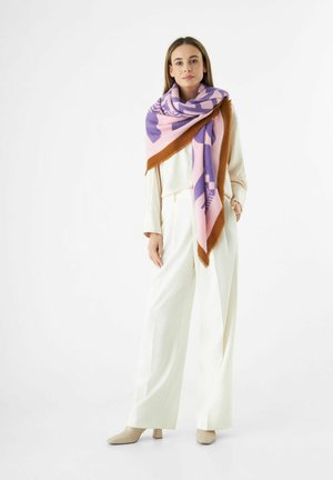 Écharpe colorée surdimensionnée dans des teintes de violet, rose et marron, avec un motif géométrique. Portée sur une tenue blanche, avec des chaussures légères.