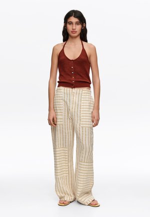 Mujer con top halter sin mangas de color óxido con botones y pantalones crema sueltos con finas rayas verticales y horizontales, de pie descalza.