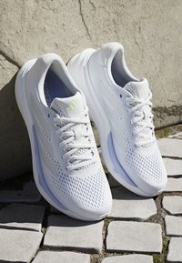 Een paar witte hardloopschoenen met mesh bovenwerk, veters en een subtiel logo, rustend op een stenen bestrating tegen een gestructureerde muur.