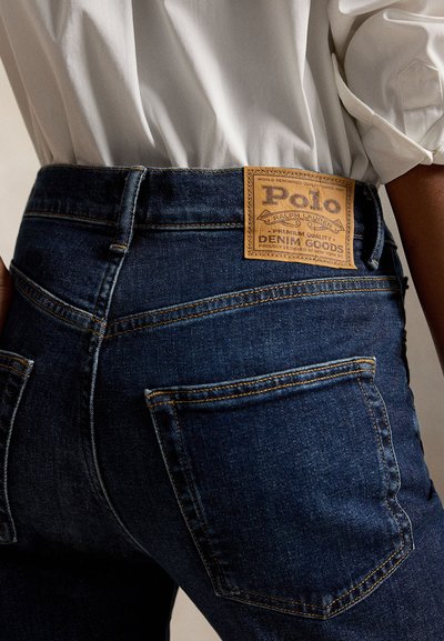 Primo piano della vita e della tasca posteriore di jeans in denim blu scuro con etichetta marrone Polo Ralph Lauren, abbinati a una camicia bianca.