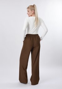 Femme blonde avec cheveux attachés en queue de cheval, portant un haut blanc à manches longues et un pantalon large marron, debout les mains dans les poches, légèrement tournée vers la gauche.