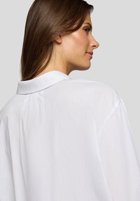 White button-up blouse featuring a classic collar, relaxed fit, and subtle texture. Includes pleat detail at the back for added style.