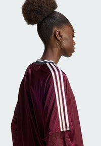 Αθλητική φανέλα χρώματος βυσσινί με λευκές ρίγες στους ώμους, διαθέτοντας υφασμάτινη υφή και μαύρη λεπτομέρεια στο γιακά.