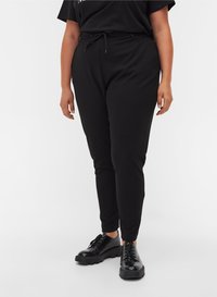 Personne portant un pantalon noir ajusté avec une taille à cordon de serrage et des chaussures à plateforme noires brillantes à lacets sur fond blanc.