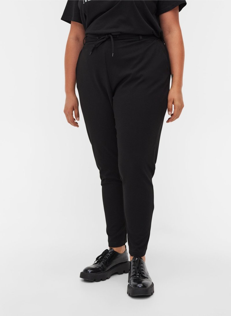 Personne portant un pantalon noir ajusté avec une taille à cordon de serrage et des chaussures à plateforme noires brillantes à lacets sur fond blanc.