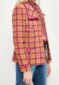 Veste en tweed orange et rose à motif à carreaux noirs, avec une finition le long du col et des poches. Associée à un chemisier rose et un jean en denim.