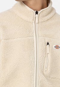 Giacca in pile beige con collo alto, cerniera frontale completa e orlo a coste. Presenta una tasca sul petto e una toppa con logo. Tessuto testurizzato e morbido.