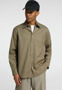 Camicia verde oliva a maniche lunghe con taschino sul petto, orli cuffi, abbinata a una maglietta bianca sotto e pantaloni khaki. Cappellino nero indossato.