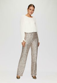 Witte geplooide lange mouwen top gecombineerd met glanzende gouden pailletten brede broek. Model draagt gouden hakken en poseert tegen een neutrale achtergrond.