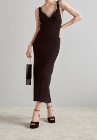 Robe noire en tricot côtelé avec un décolleté en V et des bretelles embellies, accompagnée d'un petit sac noir à franges et de talons compensés.