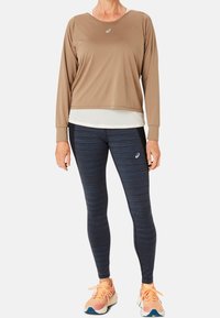 Langärmliges braunes Sportoberteil über einer weißen Schicht, kombiniert mit blauen gestreiften Leggings. Die Schuhe sind pfirsichfarben und beige mit blauen Akzenten.