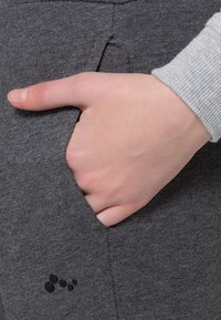 Pantalones jogger de felpa gris con una cintura acanalada, con una mano en el bolsillo lateral. Detalle de logo negro pequeño cerca de la parte inferior.