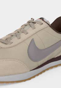 Κοντινή λήψη μπεζ αθλητικού παπουτσιού Nike με γκρι σήμα, μπεζ κορδόνια, λευκή μεσαία σόλα και καφέ εξωτερική σόλα.
