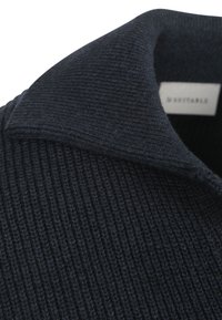 Donkerblauwe geribbelde gebreide kraag met een gladde textuur. Bevat een label met de merknaam zichtbaar bij de halslijn.