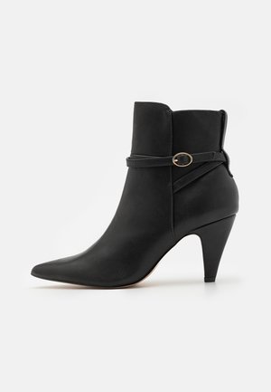 Bottine noire en matériau synthétique lisse, avec un bout pointu, un tige incurvée et une sangle décorative avec un matériel en or rond.