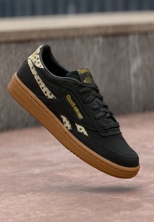 Czarny sneaker Reebok z gumową podeszwą, akcentami w panterkę i złotym brandingiem, unoszący się nad gładką różowawą powierzchnią na zewnątrz.