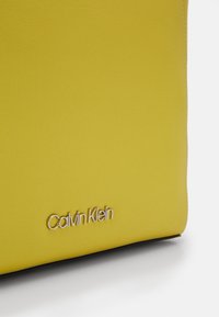 Gul læderhåndtaske med en glat overflade og et metalisk sølv "Calvin Klein" logo nederst. Minimalistisk design med afrundede hjørner.