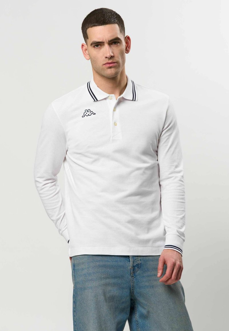 Homme aux cheveux courts et foncés portant un polo blanc à manches longues avec des bordures noires et un jean bleu clair, debout devant un fond neutre.