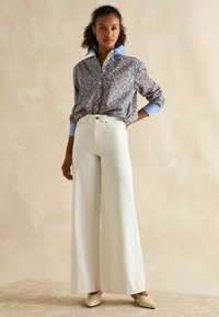 Floral patterned shirt with blue collar, paired with wide-leg white trousers. Model wearing beige heels. Plain light background.