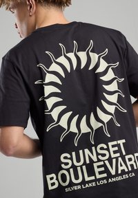 Black cotton t-shirt featuring a large white sun graphic on the back with curvy rays and the text “SUNSET BOULEVARD SILVER LAKE LOS ANGELES CA.”