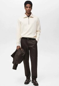 Maglione zip-up crema con colletto alto, abbinato a pantaloni marrone scuro. Il modello tiene in mano una giacca marrone coordinata. Scarpe con dettaglio strutturato.