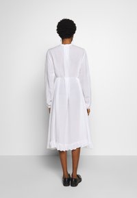 Biała sukienka z długim rękawem, z wcięciem w talii i falbanii na dole, wykonana z lekkiego materiału, z zapięciem z tyłu i marszczonymi rękawami.