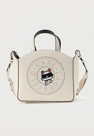 Cremefarvet håndtaske med sort kant og et sejt kattemotiv iført solbriller, centreret i et design med perforerede cirkler. Tasken har øverste hank og justerbar rem.