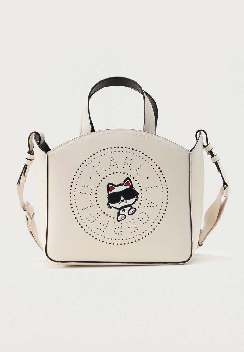 Κρεμ τσάντα χειρός με μαύρο περίγραμμα που απεικονίζει μια κουλ γάτα με γυαλιά ηλίου στο κέντρο ενός διακοσμητικού τρύπωμα σε σχήμα κύκλου, με πάνω χερούλι και ρυθμιζόμενο ιμάντα.