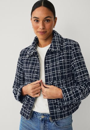 Woman with dark hair wearing hoop earrings, a navy blue and white checkered jacket over a white top, and blue jeans against a plain background.